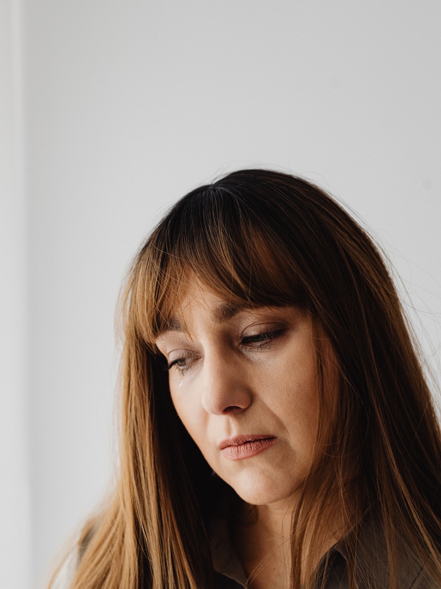 A woman with bangs against a white wall, looking down and sad.
