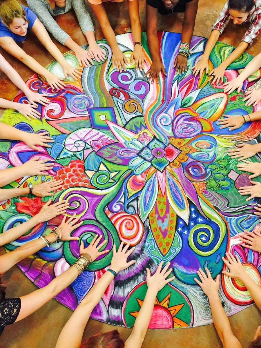 A bunch of women's hands, reaching towards the middle of a giant colourful art piece.