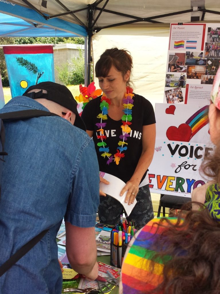 A woman with a rainbow lea around her neck and informational banners behind her while a man looks on.