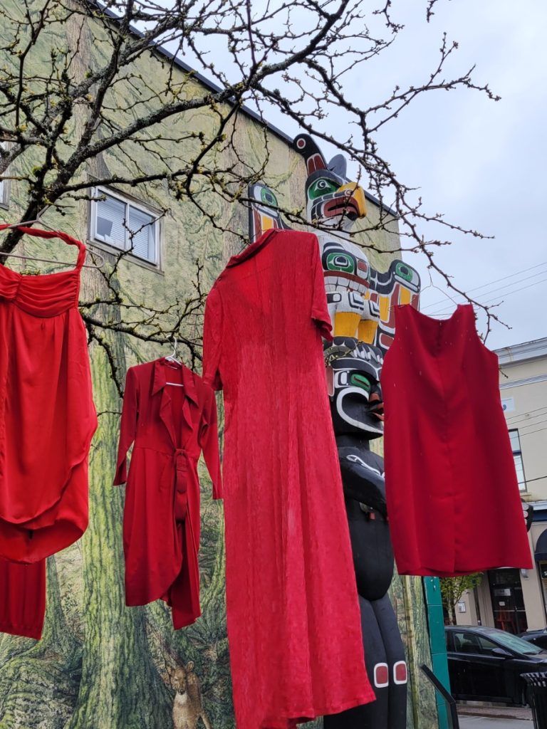 Four red dresses hanging in a tree with a First Nation's totem pole behind.