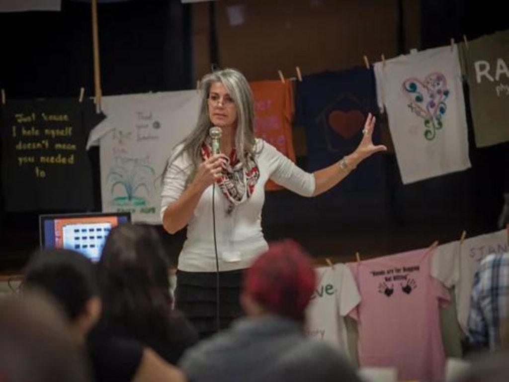 A woman standing in front of t-shirts with writing on it, with a microphone in her hand.